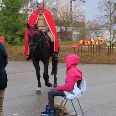 Sankt Martin ist mit seinem roten Mantel auf dem Pferd groß in der Bildmitte zu sehen. Vor ihm, mit dem Rücken zum Betrachter, sitzt ein Mädchen auf einem Stuhl. Sie spielt die Bettlerin, neben ihr liegen Lumpen.