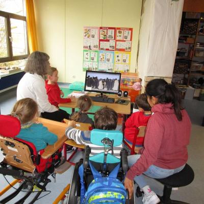 2 Frauen und 5 Erstklässler sitzen in einem Klassenzimmer vor einem PC Bildschirm, auf dem eine Kolonie Kaiserpinguine zu sehen ist. 2 Kinder sitzen im Rollstuhl bzw. Therapiestuhl, die anderen auf Stühlen.