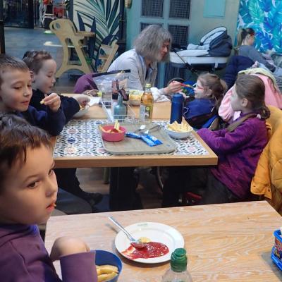 Im Restaurant der Wilhelma sitzen 3 Erstklässler links und 2 Mädchen rechts am Tisch. 4 Kinder essen Pommes, ein Mädchen hinten links bekommt von ihrer Lehrerin in der Mitte das Essen mit einem Löffel gereicht.