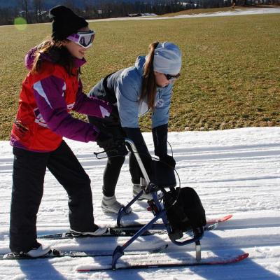 Marija läuft in der Loipe mit einem Skilator (Ski-Rollator). Frau Fecke unterstützt sie dabei, die Richtung zu halten.