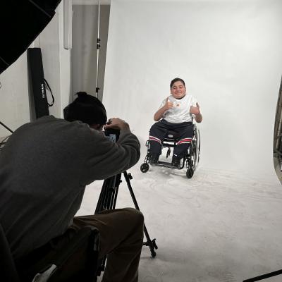 Ein Junge im Rollstuhl streckt beide Daumen hoch und lächelt in die Kamera während der Kameramann die Kamera und das Licht einstellt. 