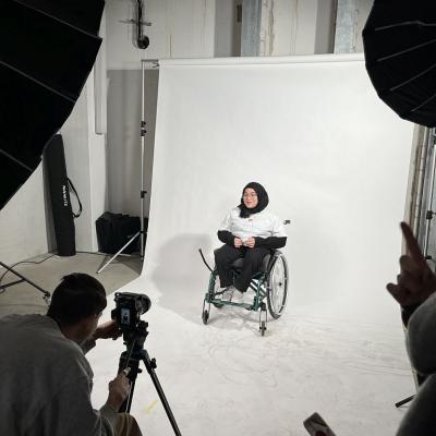 Ein Mädchen mit einem Kopftuch und einer Brille sitzt in einem Rollstuhl und probt die Gebärden vor der Kamera.