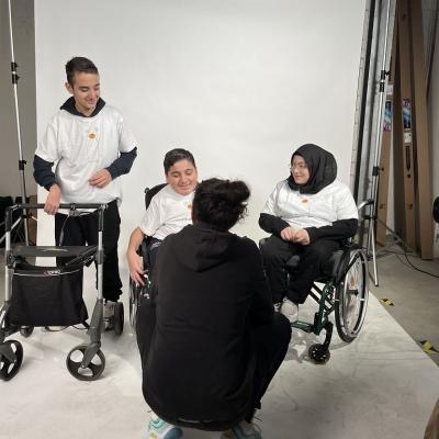Zwei Jungs und ein Mädchen werden interviewt. Die Schüler sind im Fotostudio der MHP-Riesen. Zwei Schüler sitzen in einem Rollstuhl während ein Schüler einen Rollator hat. Der Interviewer sitzt in der Hocke und nimmt das Interview mit einem Handy auf.
