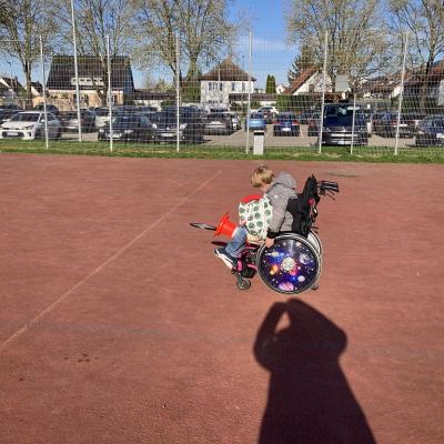 Ein Kind sitzt im Rollstuhl und hat eine Tasche mit Sportgeräten auf dem Schoß und einen Pylonen mit einem Tennisschläger zwischen den Beinen. 