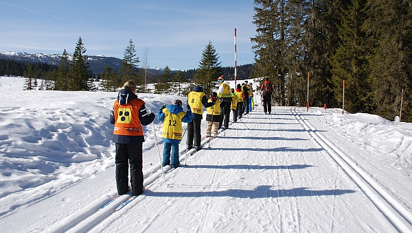In einer Loipe stehen mehrere Kinder hintereinander. Alle tragen farbige Leibchen. Die Umgebung ist schneebedeckt. Rechts beginnt ein Wald. Im Hintergrund sind schneebedeckte Berge zu sehen.