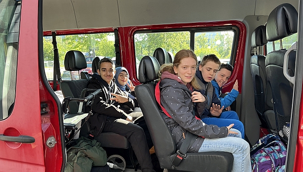 3 Jungs und 2 Mädchen in  Einem Bus und alle lachen. Im Bus sieht man auch Gepäck und einem Schulranzen und einen Rollator.