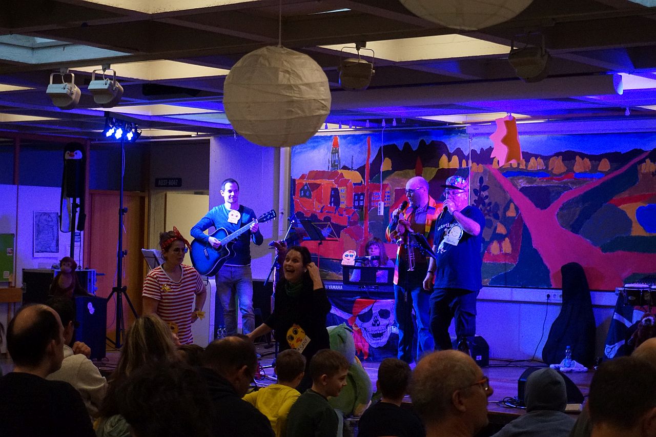 Vier Musiker sind auf der Bühne, links einer mit Gitarre, im Hintergrund eine Frau am Keyboard auf der rechten Seite zwei Sänger mit Mikrofonen. Zwei verkleidete Musikerinnen stehen vor der Bühne und animieren das Publikum 