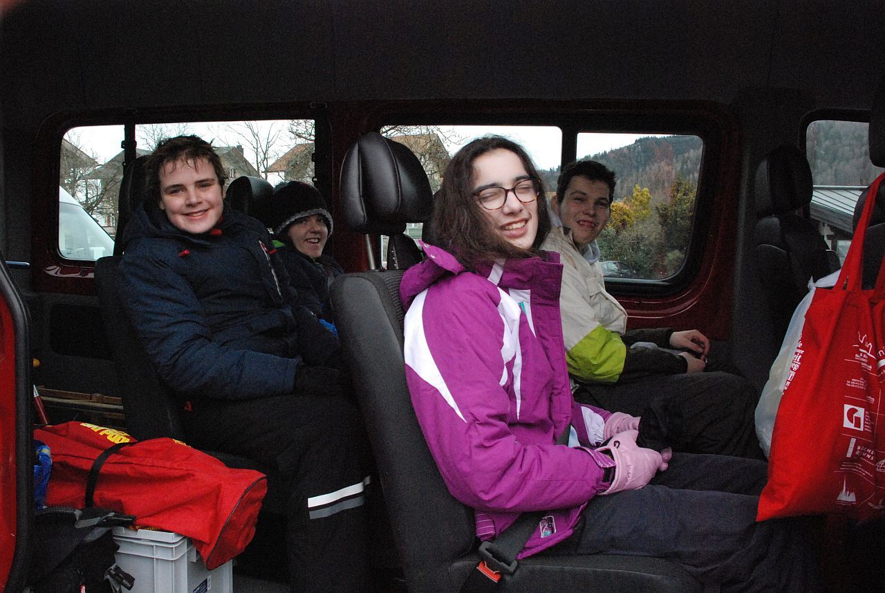 Im Schulbus sitzen vier Kinder. Auf der hinteren Rückbank sitzen Christopher und Josua und davor Marija und Julian – alle mit warmen Skiklamotten. Alle lächeln in die Kamera. Im hinteren Bereich des Busses sieht man Schlitten, einen Skisack und eine graue Kiste stehen.