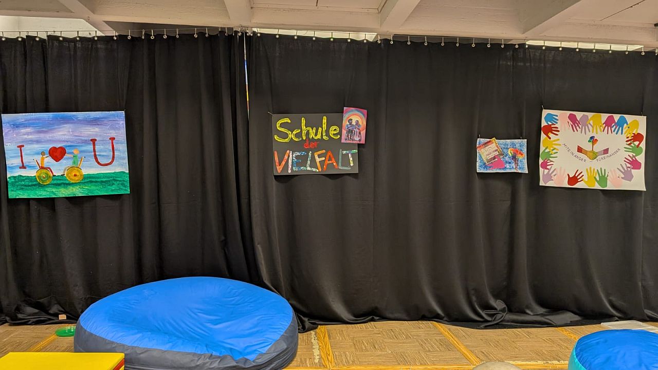 In der Aula hängen über der Bühne vier Plakate. Auf einem sieht man zwei Rollstuhlfahrende und die Botschaft „I love you“. Auf einem steht „Schule der Vielfalt“. Auf einem sind viele bunte Hände und darunter steht „Miteinander – Füreinander“.
