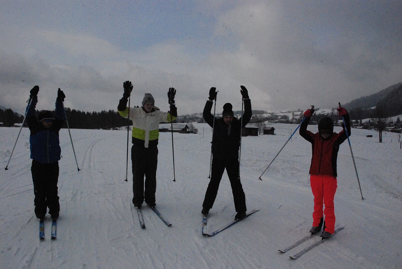 Josua, Julian, Christopher und Lara stehen auf Langlaufski im Schnee in einer Reihe und heben die Arme nach oben, wie beim Aufwärmen. Im Hintergrund sieht man viel Schnee und bewölkten Himmel.