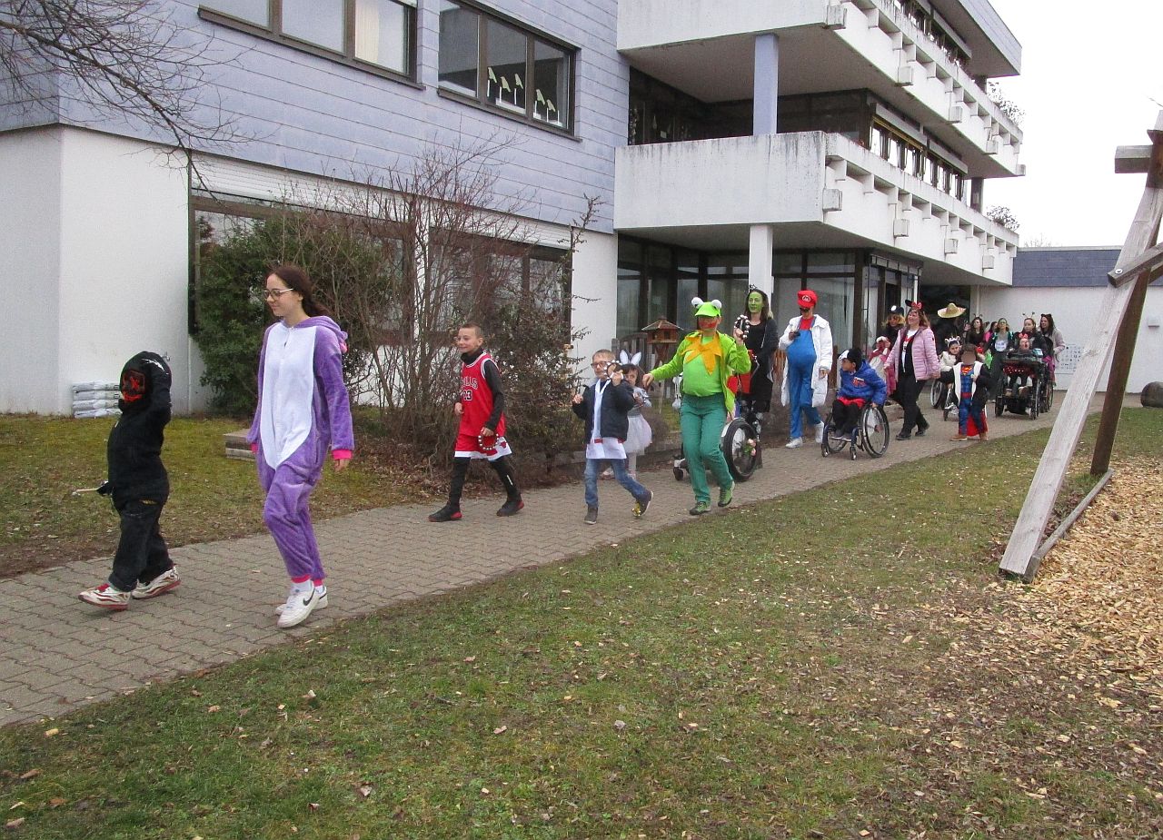 Verkleidete SchülerInnen und Mitarbeitende der Grundstufe (Klasse 1-4) draußen auf dem Weg zur Faschingsfeier, im Hintergrund der E-Bau der AHWS