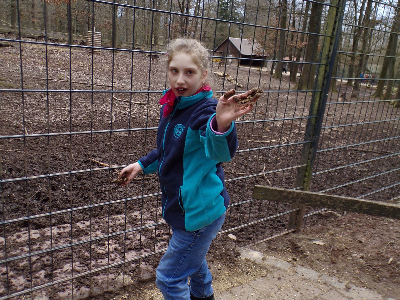 Schülerin steht vor dem Gehege am Zaun im Hintergrund sieht mal leider keine Tiere nur das Holzhaus von denen, die Schülerin zeigt ihre dreckigen Hände in die Kamera und lacht