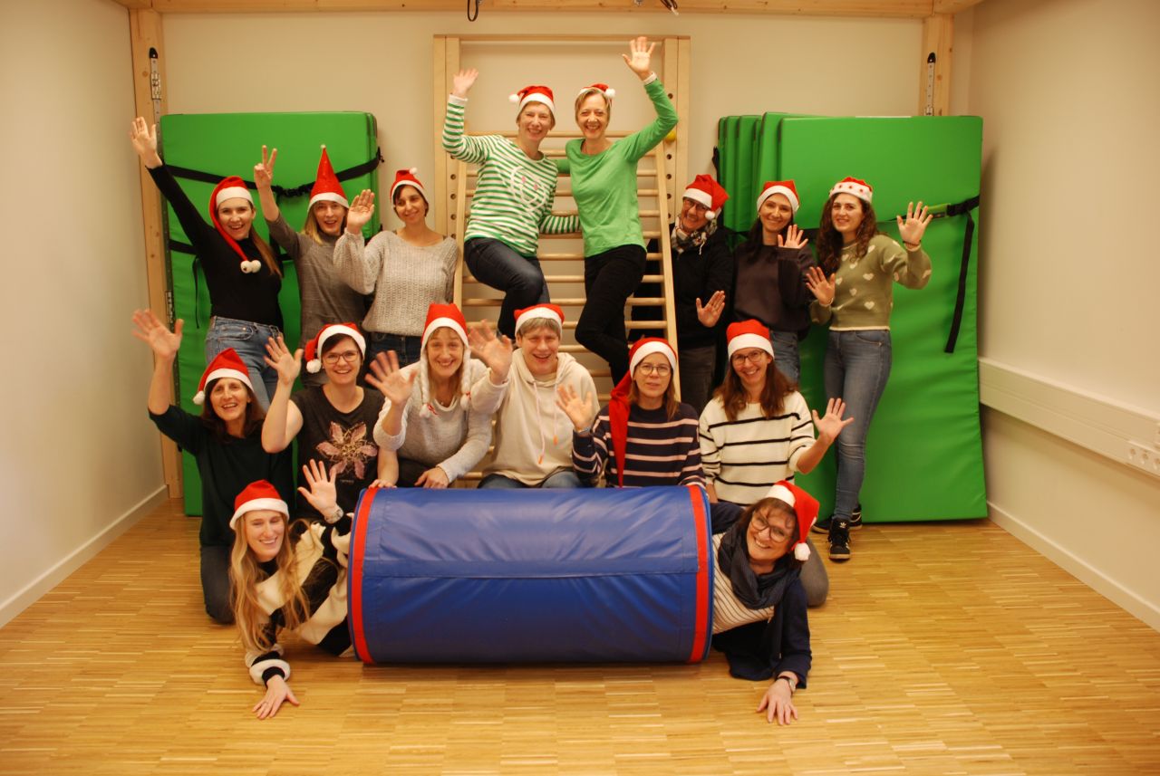 Auf dem Foto sind 16 Frauen mit Weihnachtsmützen zu sehen. Zwei Frauen stehen auf einer Sprossenwand, eine Frau schaut mit dem Oberkörper aus einer Rolle heraus und der Rest steht oder sitzt um die Rolle und die Sprossenwand herum und posiert mit einem Arm oben. 