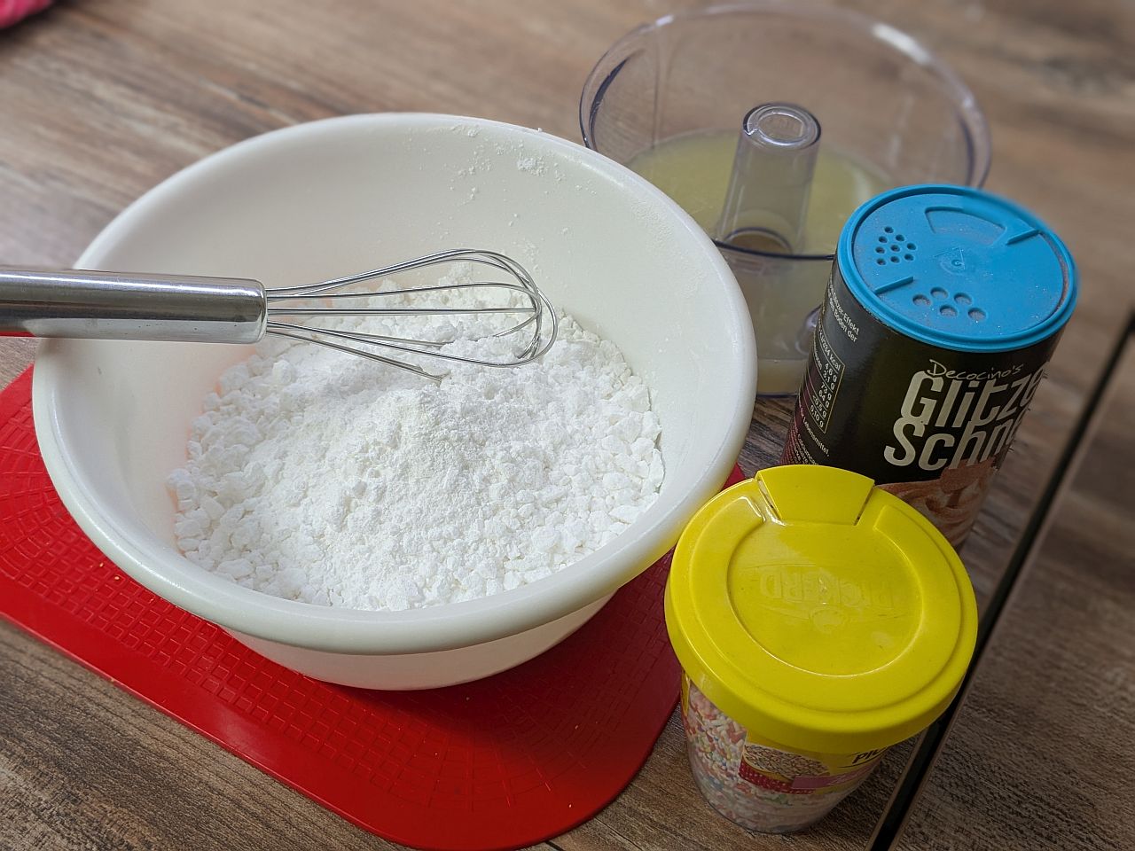 Auf dem Tisch stehen Backzutaten: Puderzucker, Zitronensaft und bunte Streusel