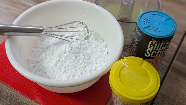 Auf dem Tisch stehen Backzutaten: Puderzucker, Zitronensaft und bunte Streusel