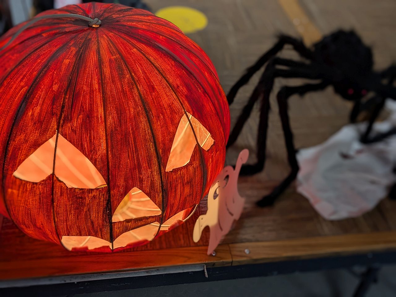 Eine leuchtende Kürbislaterne steht auf dem Boden. Neben ihr steht eine größe schwarze Spinne