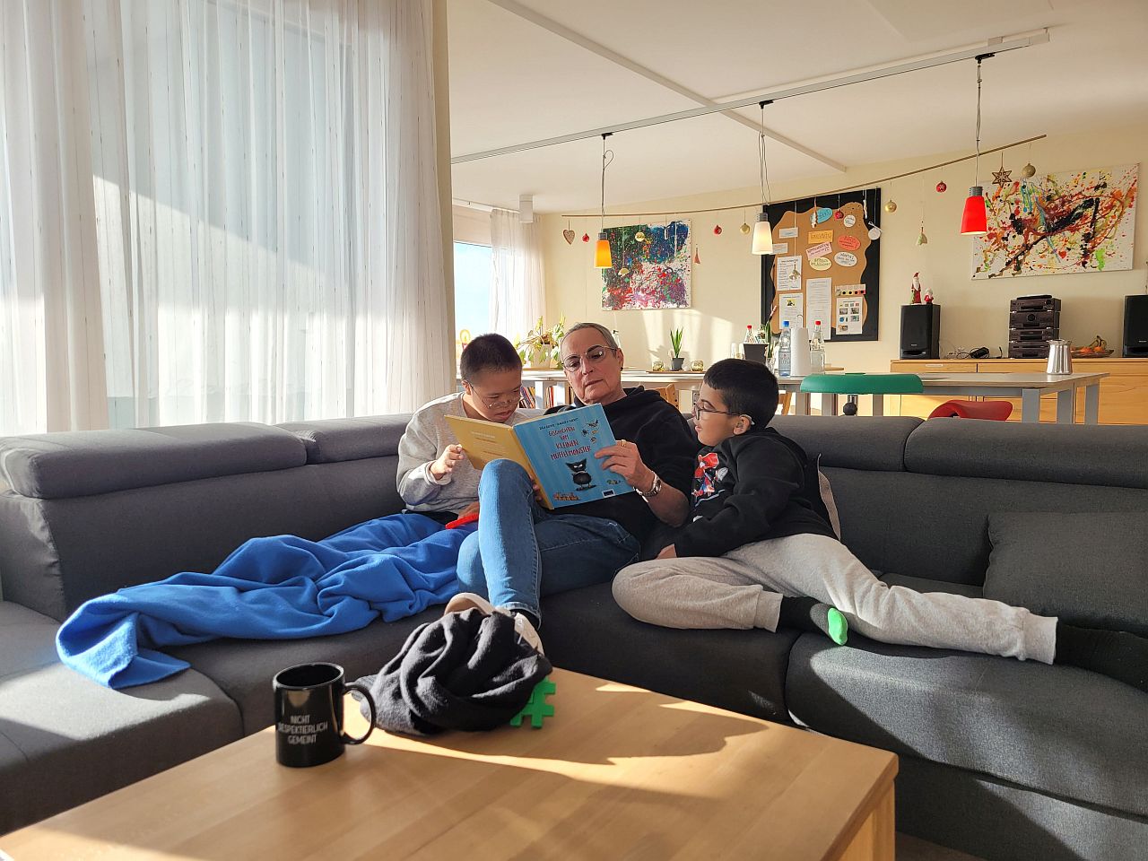 Eine Frau sitzt auf dem Sofa und liest eine Geschichte aus einem Buch vor. Links und Rechts neben ihr sitzen zwei Jungen und hören aufmerksam zu. 