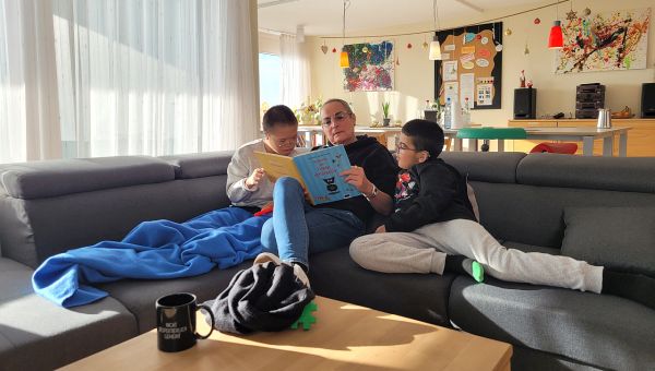 Eine Frau sitzt auf dem Sofa und liest eine Geschichte aus einem Buch vor. Links und Rechts neben ihr sitzen zwei Jungen und hören aufmerksam zu. 