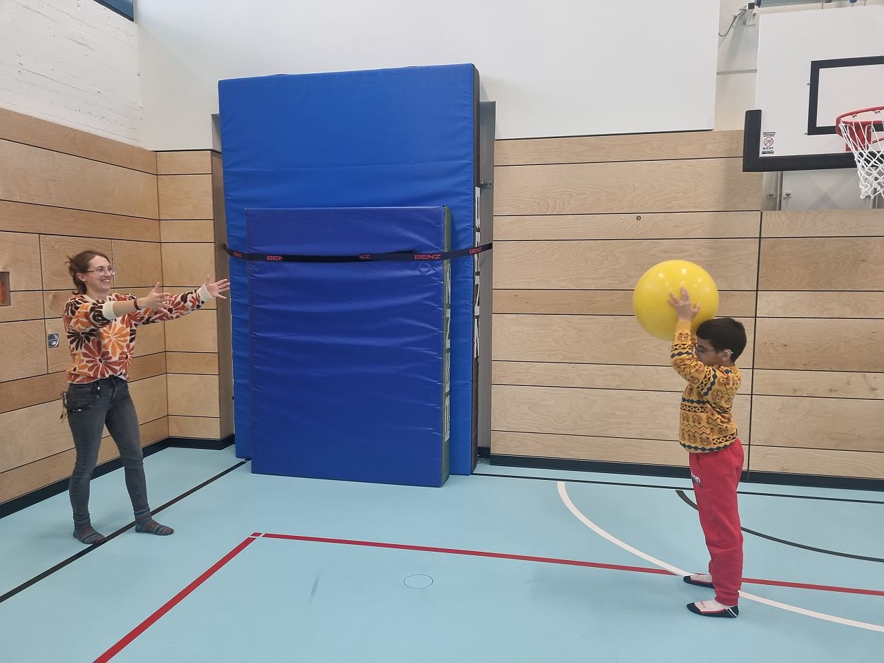 Eine Frau und ein Junge stehen sich gegenüber. Sie spielen sich einen gelben Ball zu. Sie sind in einer Sporthalle