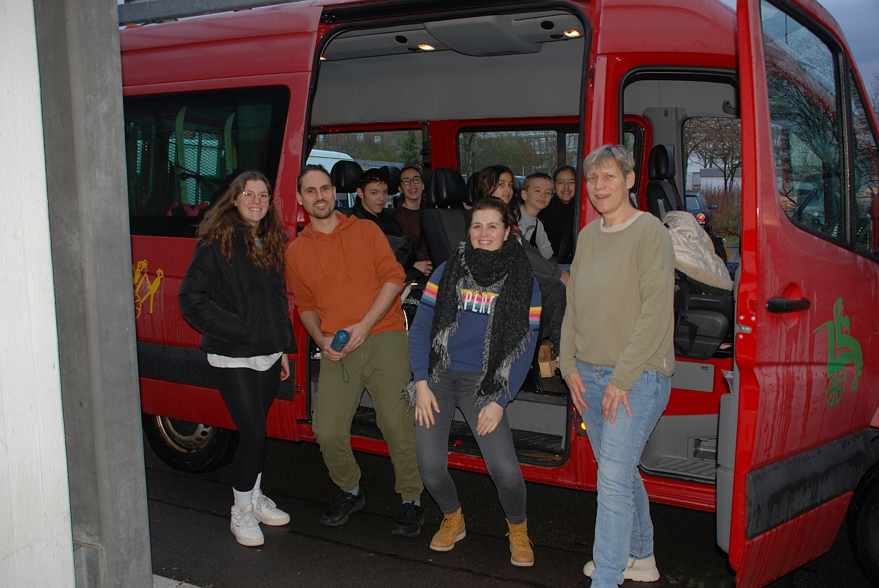 Auf den Rückbänken des roten Schulbusses sitzen Maik, Marija, Sophia, Luca und Ghofran. Davor stehen Lena, Herr Kling, Frau Fecke und Frau Gauch. Alle lächeln in die Kamera.