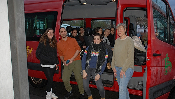 Auf den Rückbänken des roten Schulbusses sitzen Maik, Marija, Sophia, Luca und Ghofran. Davor stehen Lena, Herr Kling, Frau Fecke und Frau Gauch. Alle lächeln in die Kamera.