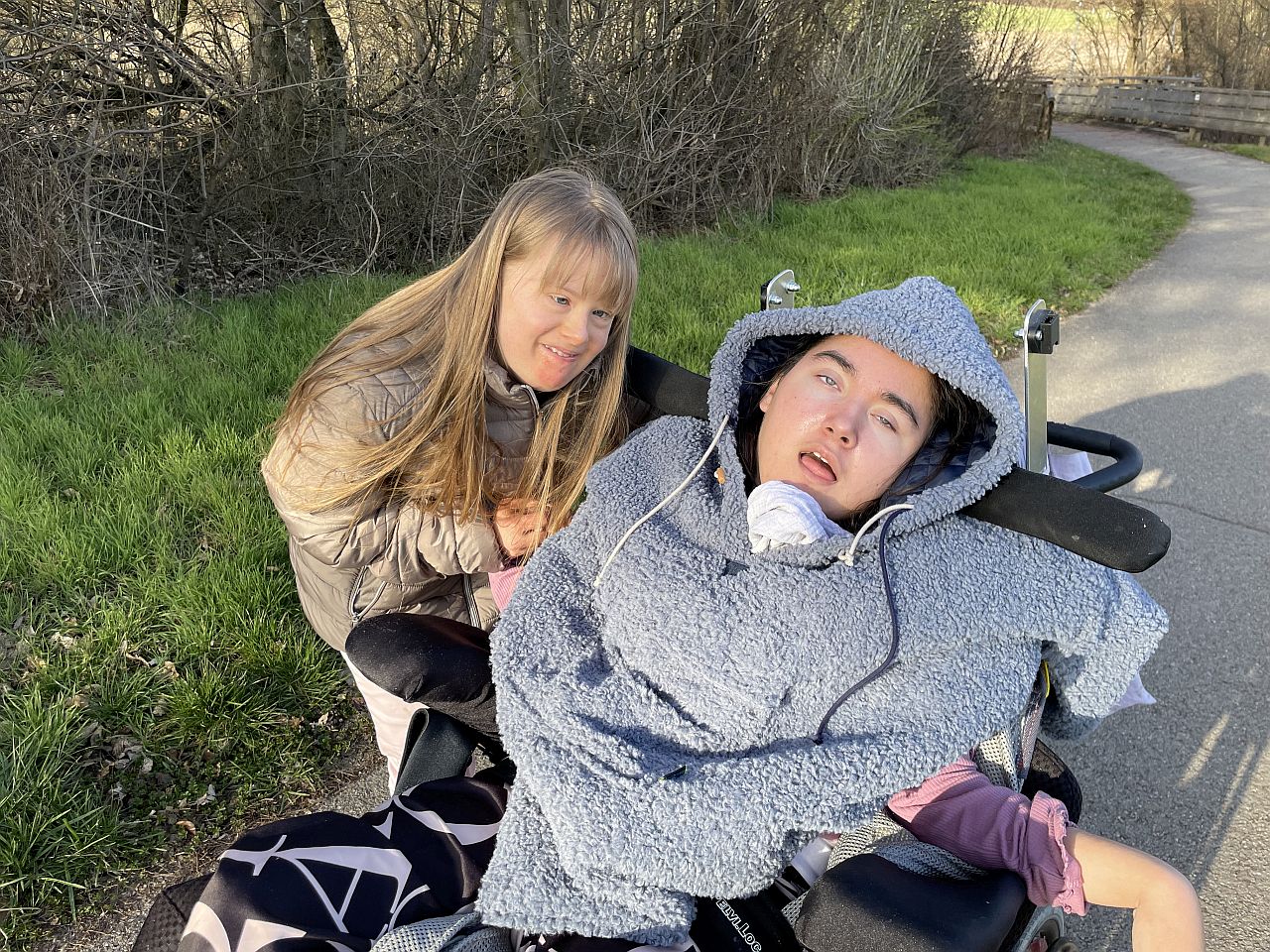 zwei junge Frauen, eine steht, eine sitzt im Rollstuhl, sind dicht beeinander und schauen in die Kamera