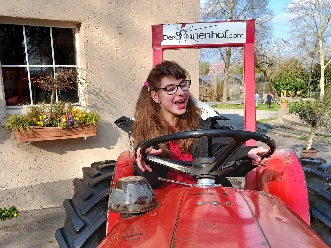 Schülerin sitzt lachend auf einem roten Traktor und hält das schwarze Lenkrad fest. Am hinteren Teil vom Traktor steht die Aufschrift "Sonnenhof "