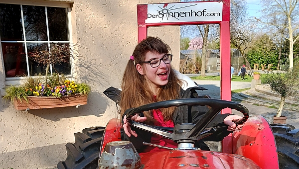 Schülerin sitzt lachend auf einem roten Traktor und hält das schwarze Lenkrad fest. Am hinteren Teil vom Traktor steht die Aufschrift "Sonnenhof "