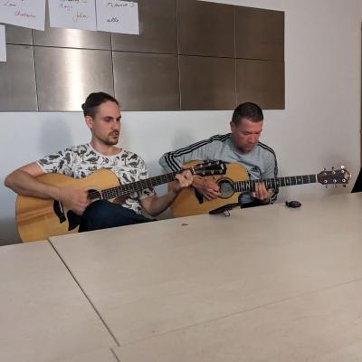 Herr Kling und Herr Schellinger sitzen am Tisch und spielen Gitarre.