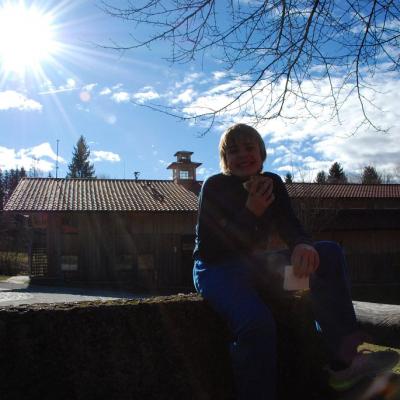 Christopher sitzt auf einer Mauer und lächelt. Im Hintergrund strahlt die Sonne am blauen Himmel.