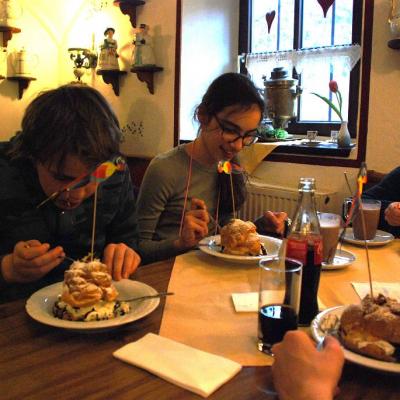 Christopher, Marija und Lara sitzen am Tisch. Rechts am Bildrand sieht man noch Florians Hände. Jeder hat einen Teller mit einem großen Windbeutel vor sich auf dem Tisch. Im Hintergrund sind ganz viele Kannen auf Regalbrettern an der Wand. 