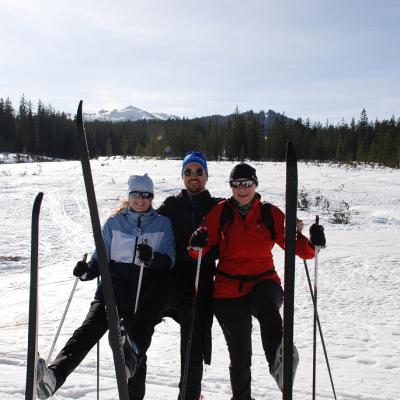 Frau Fecke, Herr Kling und Frau Gauch stehen nebeneinander. Alle haben einen Ski vor sich aufgestellt und lächeln in die Kamera. Im Hintergrund strahlt die Sonne über dunklen Bäumen und schneebedeckten Bergen.