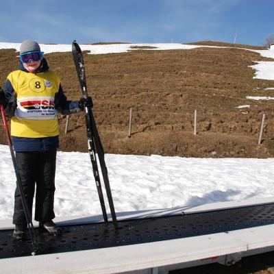 Christopher steht auf einem Förderband, das ihn nach oben befördert. Er hält seine Ski seitlich und lächelt in die Kamera.