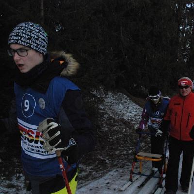 Florian läuft in der Loipe. Hinter ihm kommt Marija, begleitet von Martin von der Skiwacht. Frau Fecke läuft rechts daneben. Im Hintergrund sind dunkle Bäume.