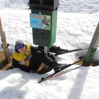 Christopher liegt auf dem Rücken im Schnee. Seine Beine mit Skiern an den Füßen gehen rechts und links an einem Hundekot-Eimer vorbei. Er grinst in die Kamera.