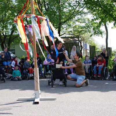 Der Maibaum steht im Vordergrund. Dahinter spricht Aziz in ein Mikrofon, das von Herrn Kling gehalten wird.