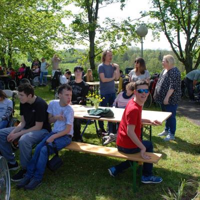Im Schatten der Bäume stehen einige Bierbänke. Daran sitzen Schüler:innen.