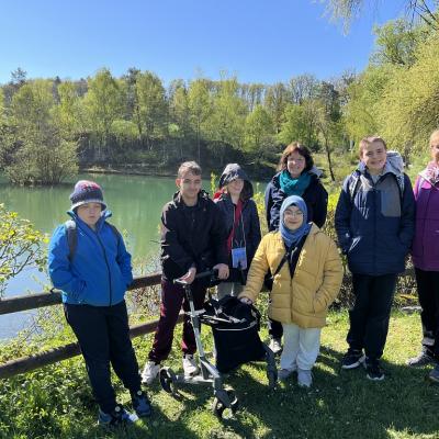 Die Klasse ist auf einem Ausflug. Sie stehen nebeneinander für ein Gruppenfoto. Hinter ihnen ist ein Holzzaun und im Hintergrund ist ein klarer See zu sehen
