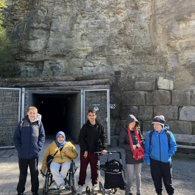 Hier seht ihr 3 Jungs und 2 Mädchen. Die Gruppe steht vor einer Höhle. Ein Mädchen mit einer gelben Jacke und einer Brille lacht.