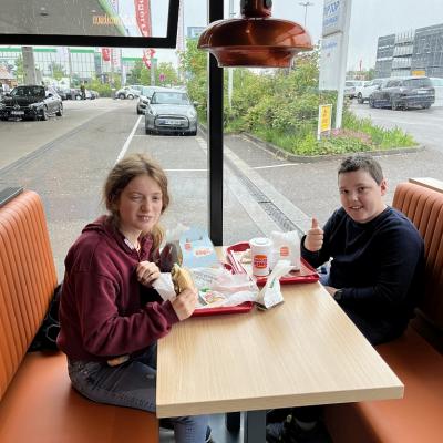 zwei Kinder im Burger King sitzen. Das Mädchen isst einen Hamburger. Der Junge hat Cola auf seinem Tablett stehen.  Im Hintergrund auf der Tankstelle stehen viele Autos auf den Parkplätzen.