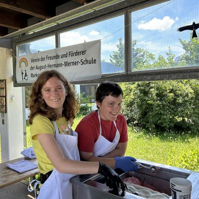 Eine Schülerin und eine Schülerin geben das Eis aus. Sie tragen Schürzen und Handschuhe und haben Eislöffel in der Hand. Im Hintergrund ist das Banner des Fördervereins zu sehen. 