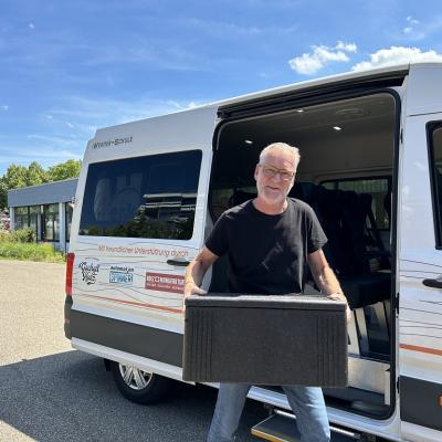 Herr Zimmermann hält eine schwarze Thermobox in beiden Händen. Er steht vor der geöffneten Tür des weißen Schulbusses des AHWerk.