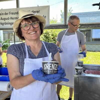 Frau Jansen trägt Handschuhe. Schürze und einen Hut. Sie hält einen Eisbecher vom Eis Café Lorenzo in der Hand und lacht. Im Hintergrund sind Erwachsen und Kinder.