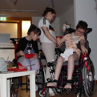 Eine Rollstuhlfahrerin schaut zu wie sich eine andere Rollstuhlfahrerin richtig hinsetzt , eine stehende Betreuerin schaut ob es richtig klappt