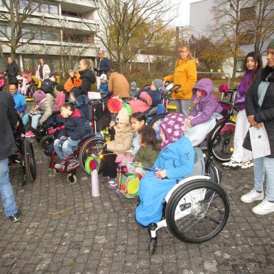 Schülerinnen und Schüler im Grundschulalter mit Laternen sitzen in Rollstühlen oder stehen mit Mitarbeitenden der Schule nach links gewandt auf dem Schulhof. Im Hintergrund ist das Internatsgebäude zu sehen.