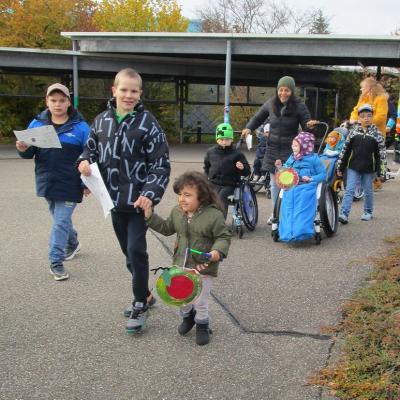 Kinder und Erwachsene ziehen mit Laternen über den Schulhof. Ein größerer Junge hält ein kleines Mädchen an der Hand.