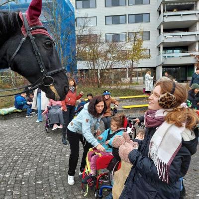 Der große schwarze Pferdekopf kommt von links ins Bild, eine Jugendliche ist rechts daneben strahlend zu sehen. Im Hintergrund weitere Kinder, zum Teil in Rollstühlen, mit Erwachsenen und das Internatsgebäude.