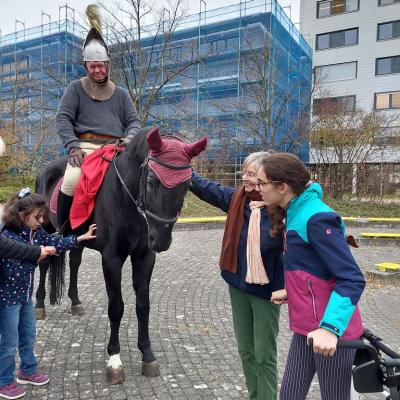 Sankt Martin auf dem Pferd links im Bild, im Hintergrund das blau eingerüstete Schulgebäude. Ein Mädchen streichelt links das Pferd, ein weiteres tritt mit seiner Lehrerin von rechts ans Pferd heran.