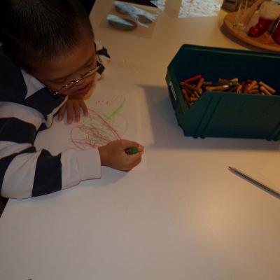 Eine Junge sitzt an einem Tisch. Vor ihm ist ein Blatt Papier. Dieses bemalt er mit verschiedenen Wachsmalstiften. Auf dem Papier ist ein buntes durcheinander zu sehen. 
