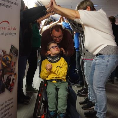 Ein Mann schiebt einen Jungen im Rollstuhl durch einen Tunnel aus Händen, der von anderen Zuschauern gebildet wird. Rechts neben dem Händetunnel ist das Plakat des Fördervereins zu sehen.